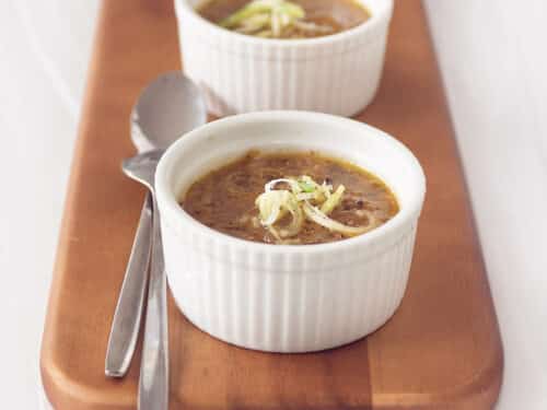 A square photo with two bowls filled with leek soup with cabbage. Two spoons next to it on a wooden board. A white background.