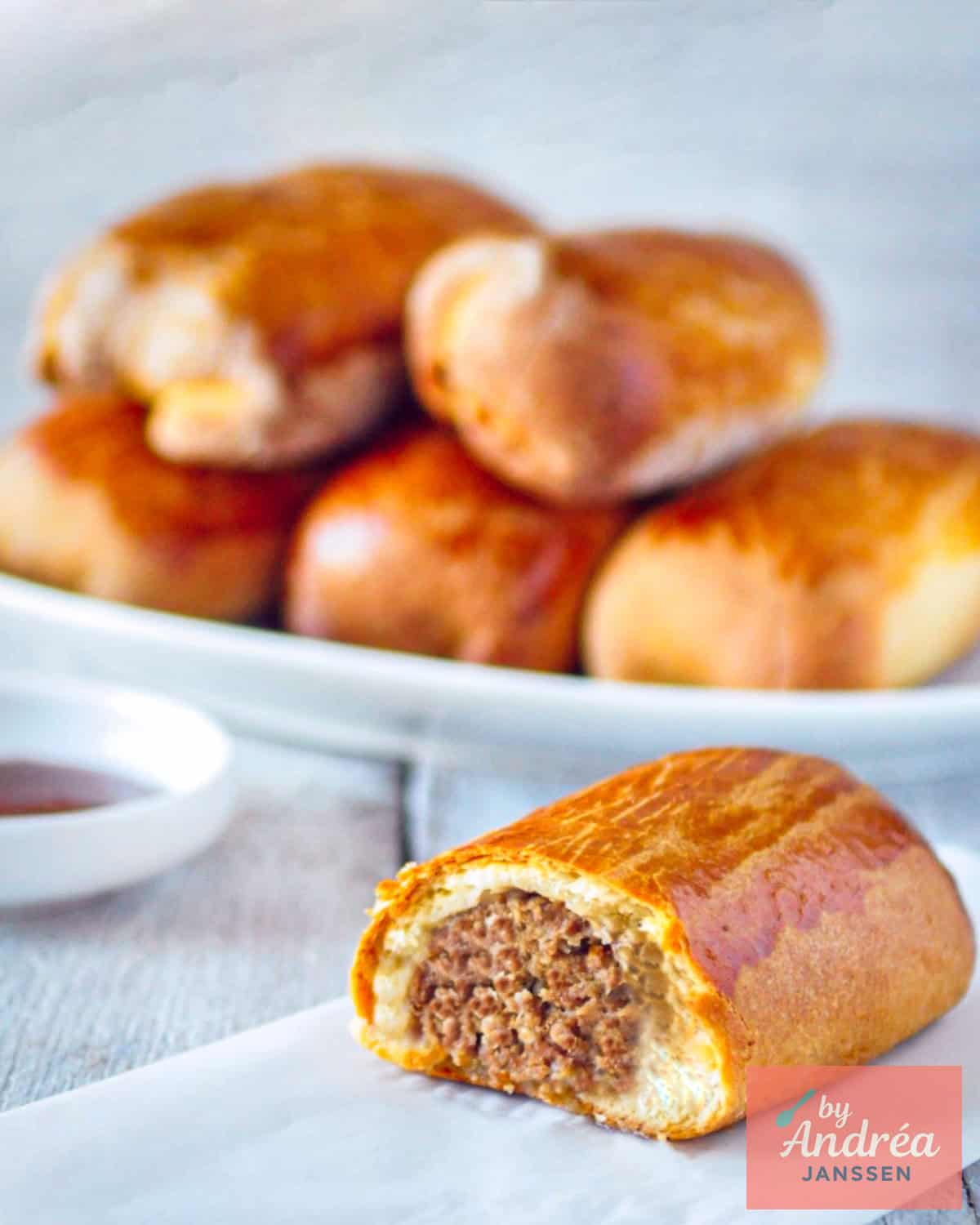 A plate with Dutch sausage rolls. In front a worstenbroodje cut in half, filliing is visible.