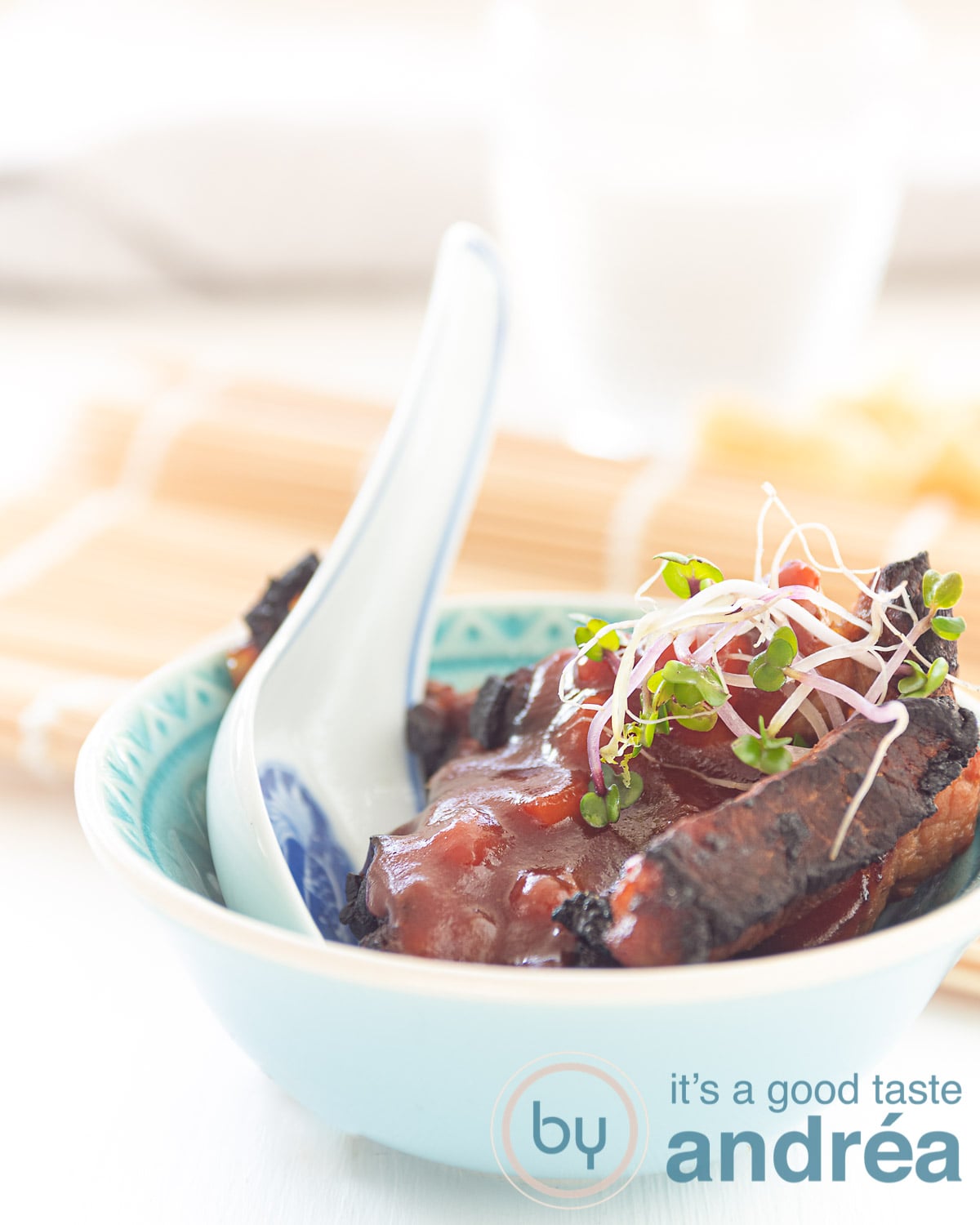 A blue dish with Babi Pangang, sweet and sour sauce and some cress on top. A white blue spoon sticks in the dish. In the background a bamboo mat
