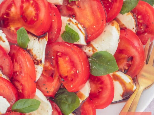 A square photo of sliced   tomatoes, basil, mozzarella and balsamic vinegar on a white plate.