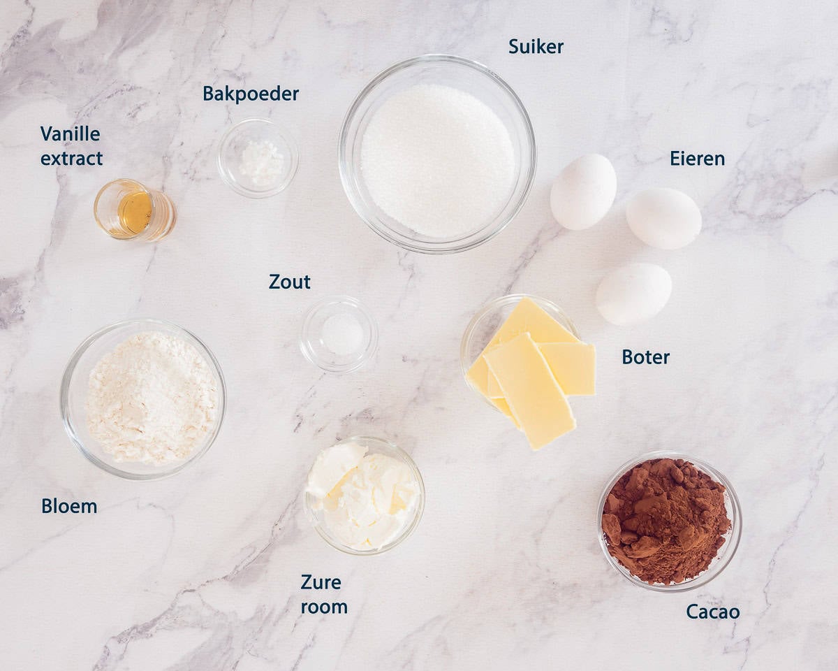 Ingredienten chocolade cupcakes op een wit marmeren ondergrond met tekstbeschrijving.