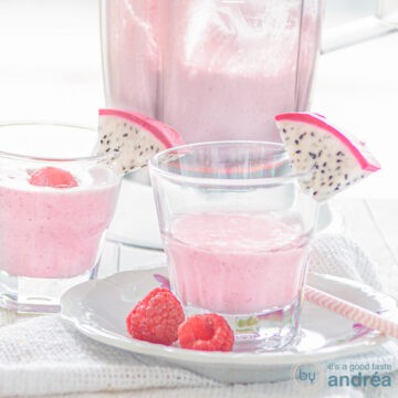 A square photo with a white plate on top of which a glass filled with dragon fruit smoothie with raspberries and a slice of dragon fruit. A white background