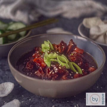 A square picture with a grey bowl filled with slow cooker mongolian beef with spring onion garnish and some prawn crackers.