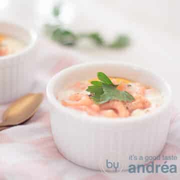 A square photo with a white dish filled with an egg shell with shrimps and cheese. Standing on a pink white tea towel. In the background a sprig of parsley