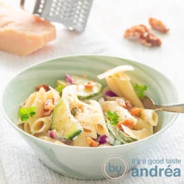 A square photo with a green bowl with penne, walnuts, zucchini and Parmesan cheese on a white background. A piece of parmesan cheese and walnuts in the background