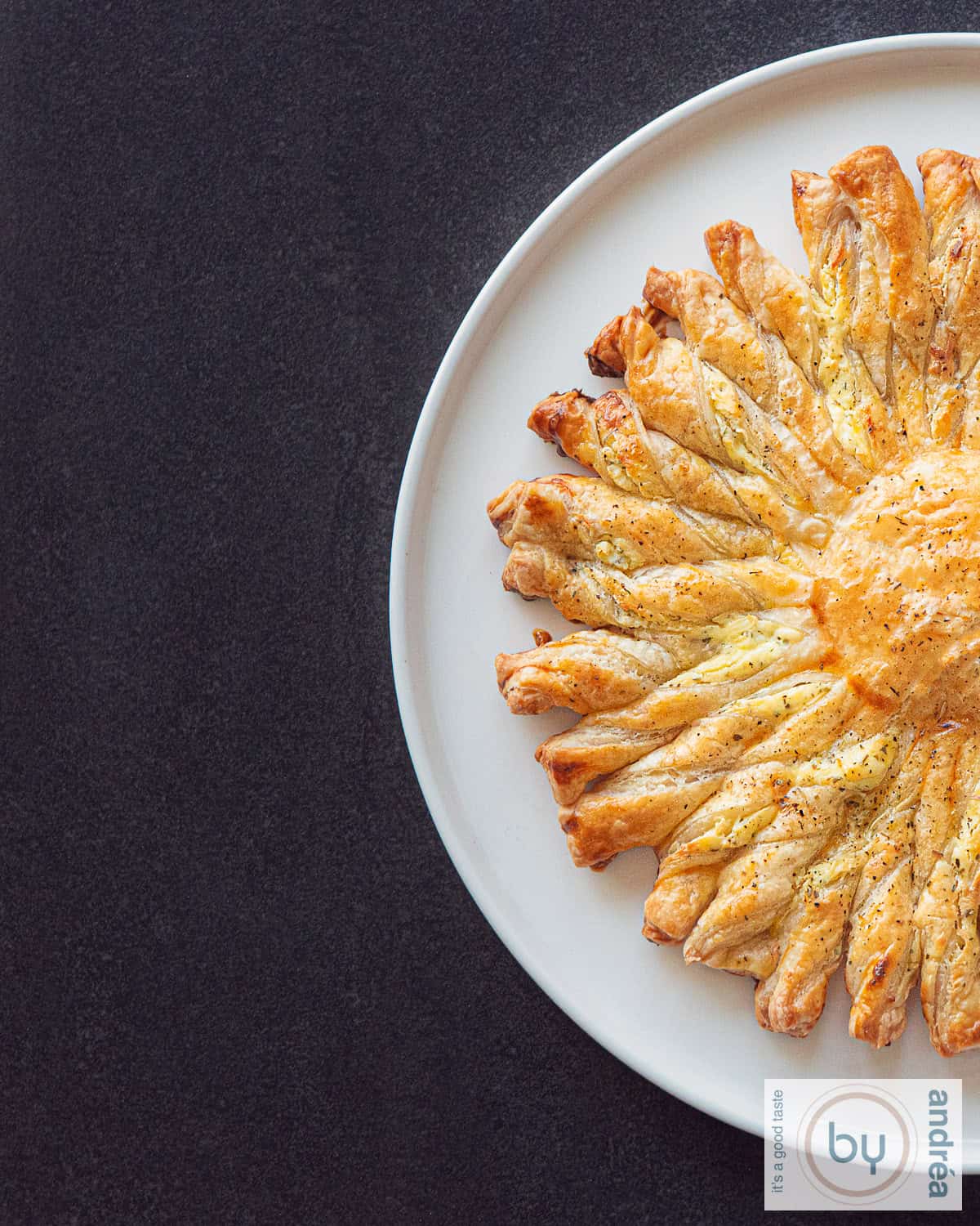 Een hoogtefoto met een donkere ondergrond, een wit bord en een halve tarte soleil met Boursin, bladerdeeghapjes die makkelijk te maken zijn er bovenop.