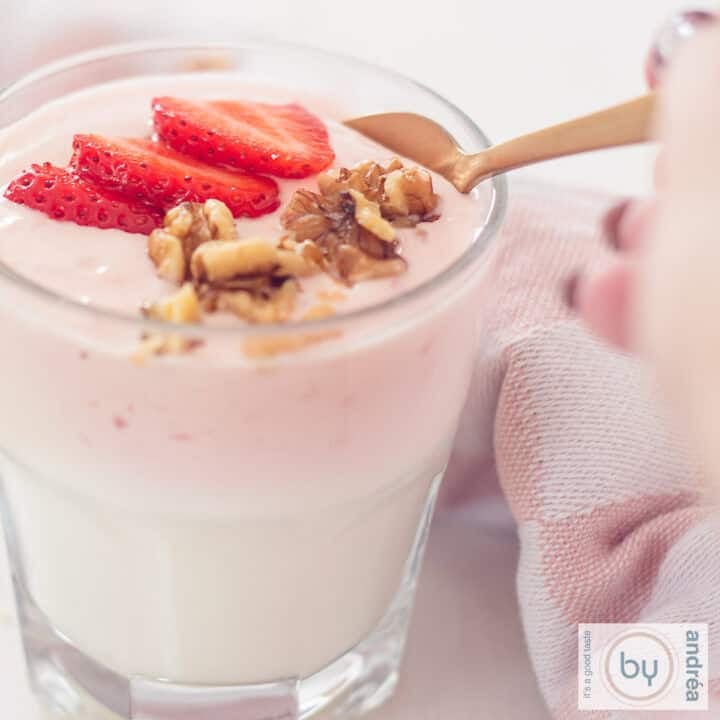 A square photo of a glass filled with homemade strawberry yogurt, walnuts and fresh strawberries. A golden spoon takes a scoop.
