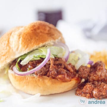 A square photo of a sandwich topped with minced meat in a delicious sauce, sloppy joe, with lettuce and red onion.