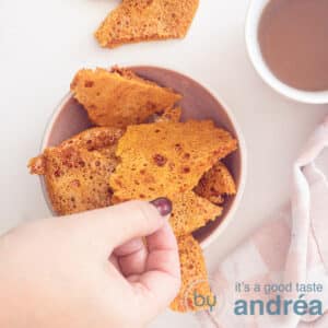 A square photo with a pink tray with honeycomb pieces in the middle. A hand grabs a piece. Top right a mug with tea.