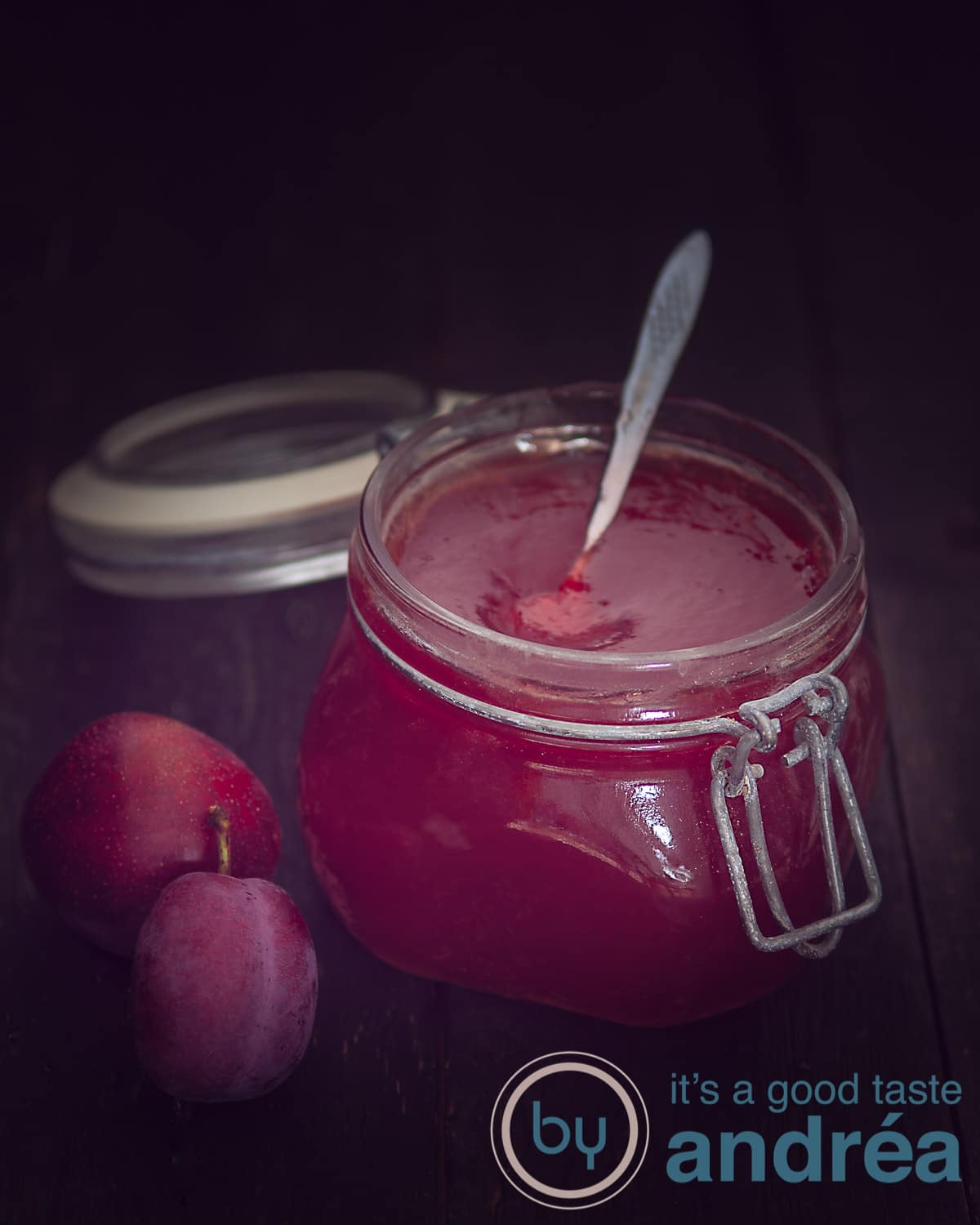 A jar of plum jam with vanilla on a dark background. Two plums on the left