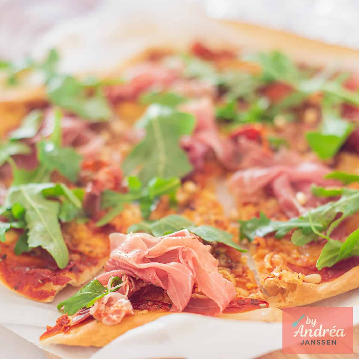 A square picture with a pizza with parma ham, arugula and pine nuts.