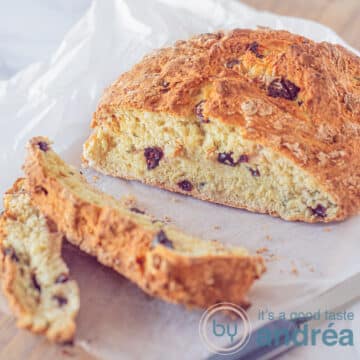 A square photo of a soda bread on a white parchment paper on a wooden board. two slices have been cut off and they are in the front left.