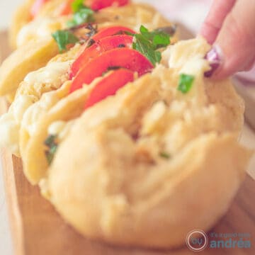 A square photo with part of a stuffed baguette filled with tomato, mozzarella and basil. A hand grabs a slice of baguette.