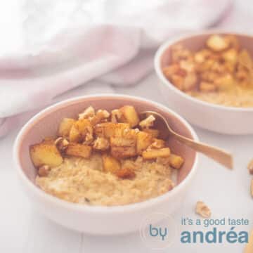 A square photo with a pink bowl in the foreground with oatmeal, apple, walnuts and honey. A golden spoon sticks into it. In the back right a second bowl. A white background with a pink and white tea towel at the top left.