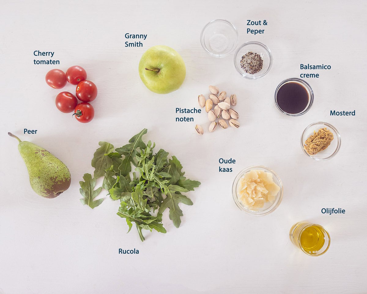 Ingredienten Rucola Salade Met Appel Peer En Pistachenoten op een witte ondergrond met tekstbeschrijving.