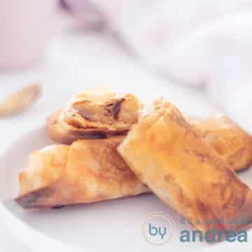 A square photograph of a white plate with a filo pastry apple turnover and two halves on top. The filling is visible.