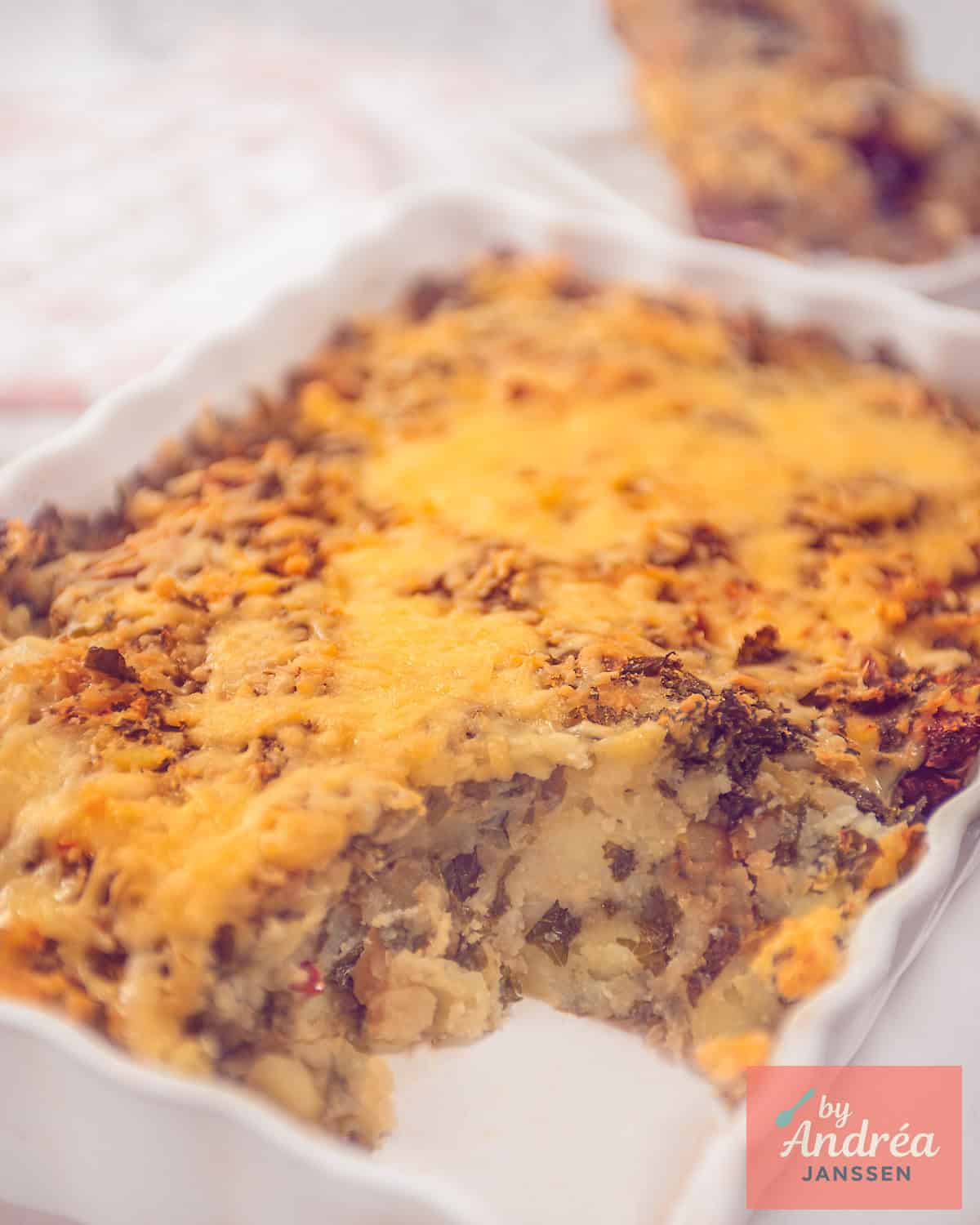 A white oven dish with a kale mash with sun-dried tomatoes under a roof of melted cheese.