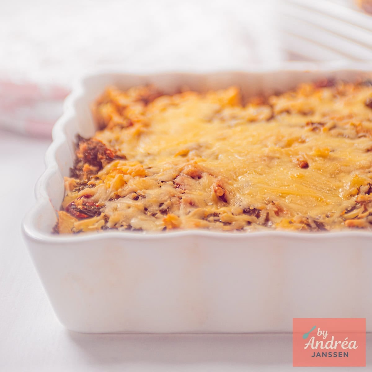 A square photo showing part of a white baking dish filled with a gratin of kale mash, cheese, and tomato