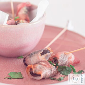 A square photo of a pink plate with a bowl filled with dates filled with blue cheese and a bacon wrap