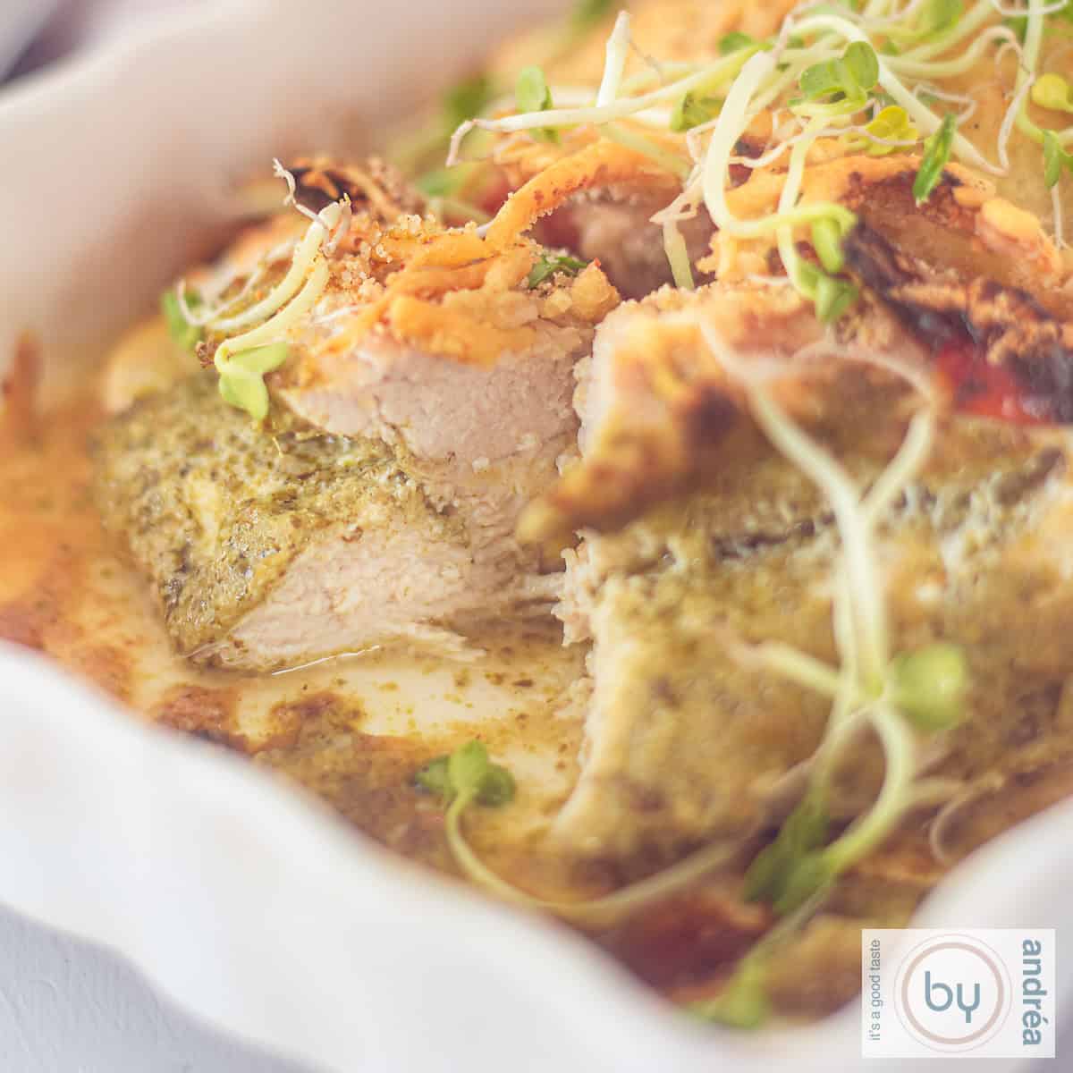 A square photo of a white baking dish, portion, chicken fillet with pesto and cheese