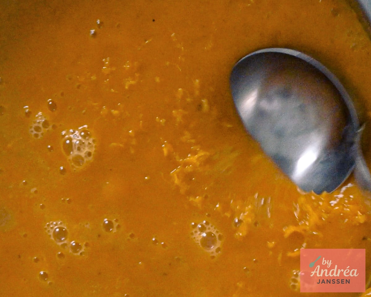 Sinaasappelrasp in oranje pompoensoep met een metalen lepel in de pan.