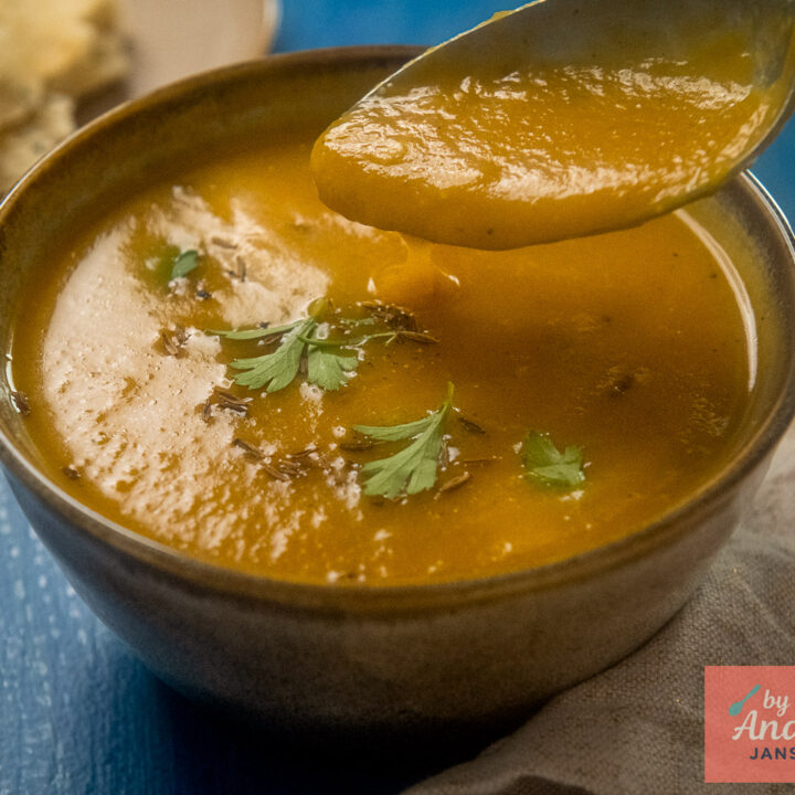 A spoonful of smooth pumpkin soup above a brown bowl of soup garnished with green herbs and cumin seeds.