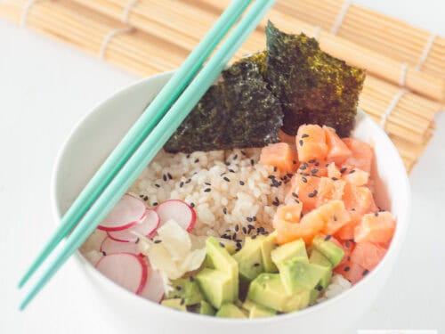 A square photo of a white bowl with sushi rice, radish, avocado, smoked salmon, and seaweed. Two blue chopsticks and a wicker mat decorate the table.