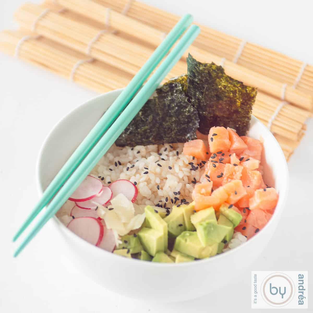 A square photo of a white bowl with sushi rice, radish, avocado, smoked salmon, and seaweed. Two blue chopsticks and a wicker mat decorate the table.