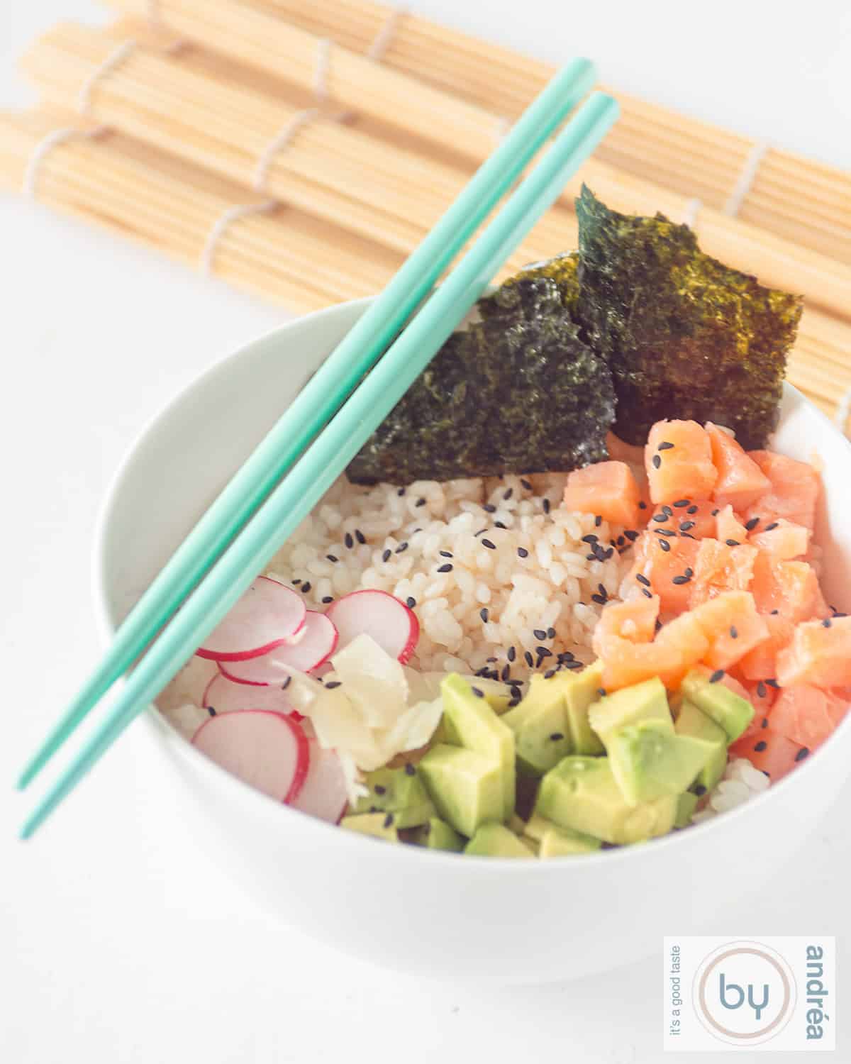 Een witte achtergrond met een witte kom gevuld met gerookte zalm, sushi, avocado, radijs en nori. Twee blauwe eetstokjes liggen op de kom.
