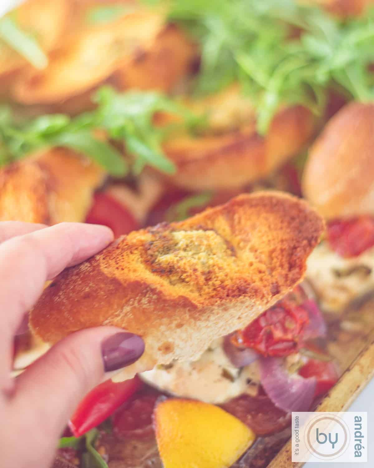 A traybake of chicken, arugula, lemon, baguette, and tomato. A hand takes a piece of baguette.