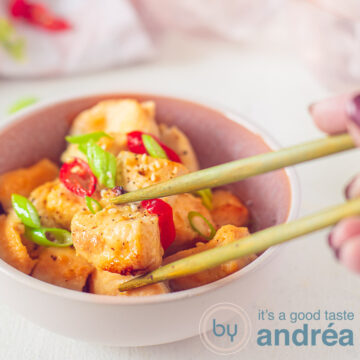A square photo with a pink dish with honey ginger chicken, spring onion and pepper. Two chopsticks grab a chicken cube. In the background a pink and white tea towel.