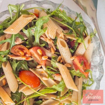 A square photo of a glass bowl with penne, cherry tomatoes, arugula, mackerel, and tapenade