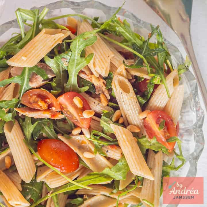 A square photo of a glass bowl with penne, cherry tomatoes, arugula, mackerel, and tapenade