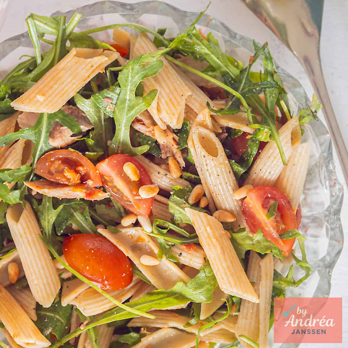 A square photo of a glass bowl with penne, cherry tomatoes, arugula, mackerel, and tapenade