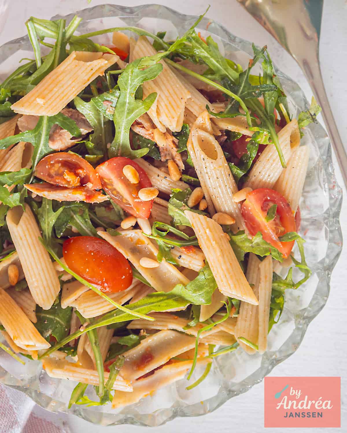 Een glazen schaal van bovenaf gefotografeerd met makreel, penne, rucola, pijnboompitten en tomaatjes