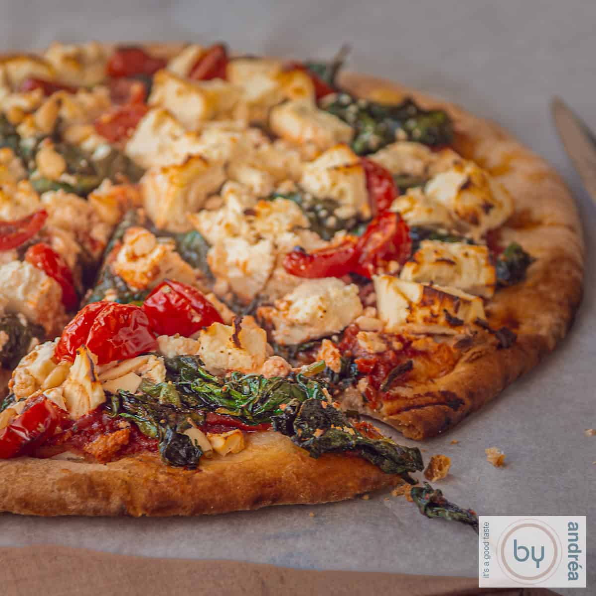 A square photo of a pizza, from which a slice has been cut, on white baking paper. The pizza is topped with feta, tomato, spinach, and pine nuts.