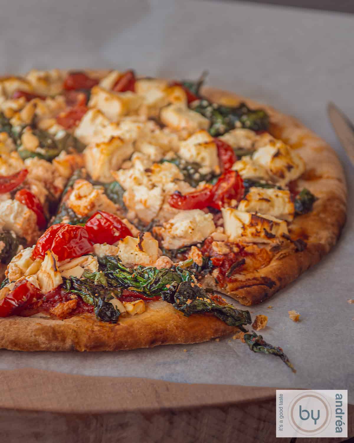 Een deel van een pizza met feta, spinazie, tomaat en pijnboompitten op een houten plank op bakpapier.