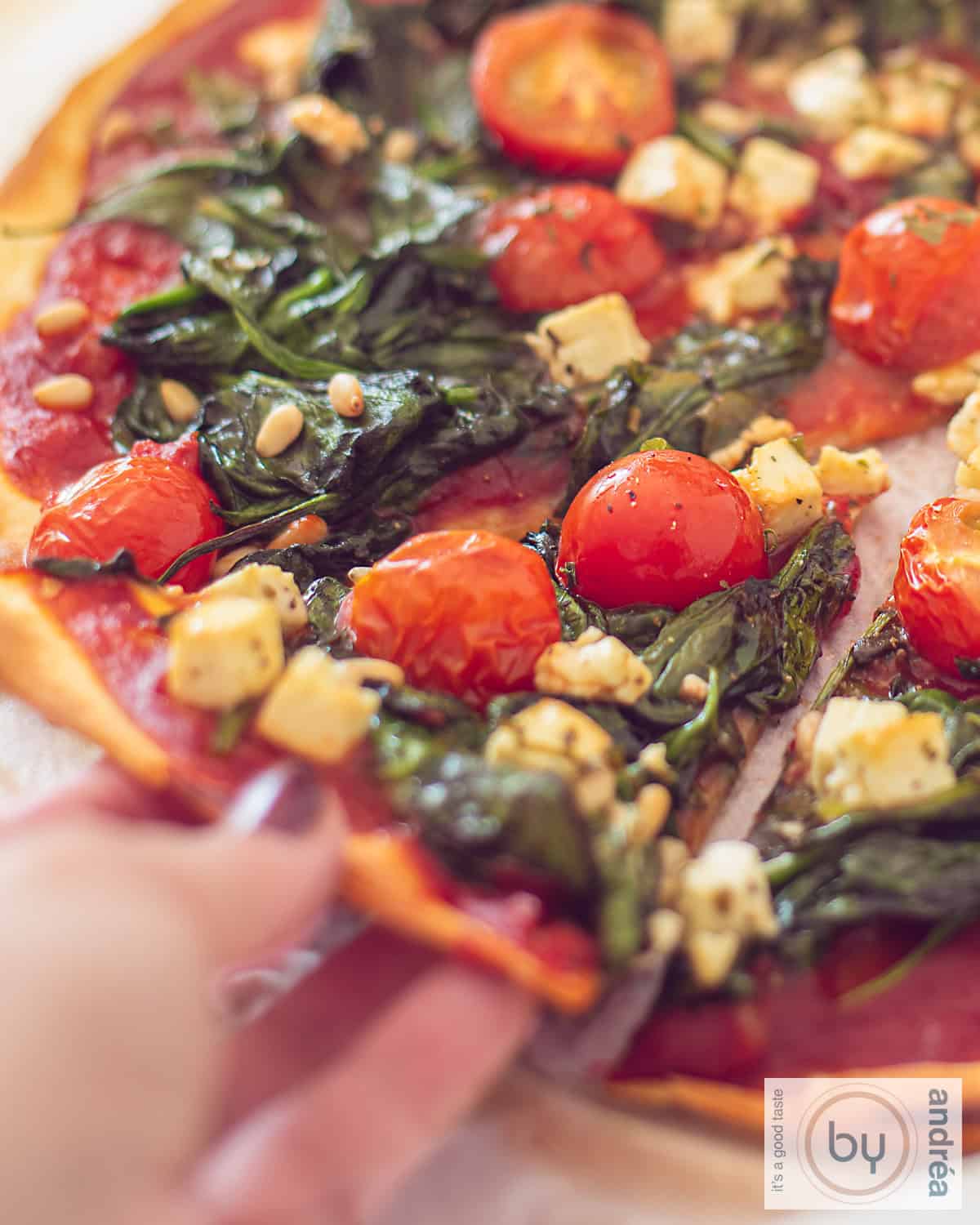 Een deel van een pizza met spinazie, feta, pijnboompitten en tomaatjes. Een punt wordt er uit gepakt.