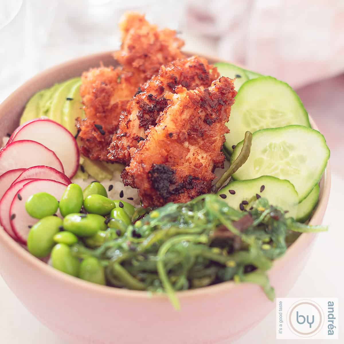 Square photo of a pink bowl with sushi bowl ingredients: crispy sriracha chicken, seaweed salad, edamame beans, radish, and cucumber.