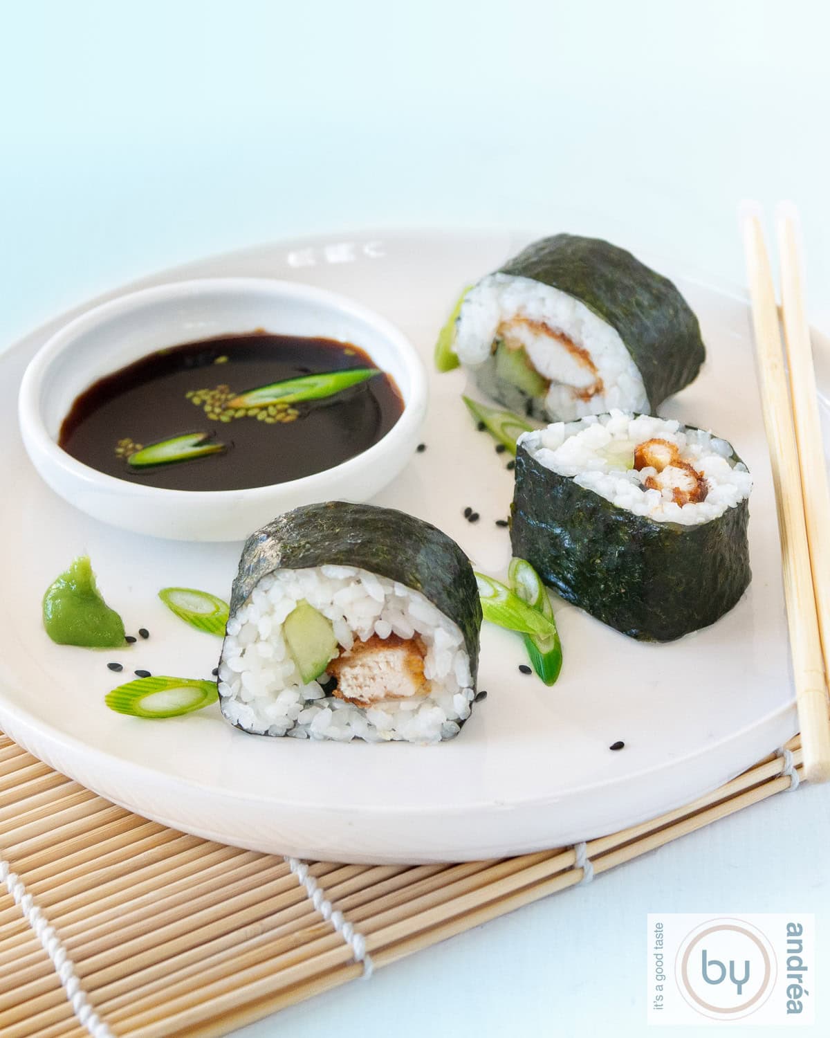 Een foto met een wit bord met drie rollen sushi met krokante kip en komkommer meet sojasaus. Een blauwe achtergrond.