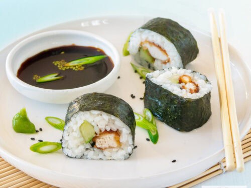 A square photo of 3 rolls of sushi with chicken and cucumber and soy sauce on a white plate.