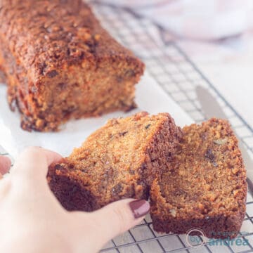 A square photo with a cake with carrots, walnuts and raisins on a baking paper on a rack. Two slices cut off. A hand grabs the top slice.
