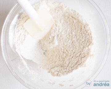 A glass bowl where the dry ingredients for the carrot cake are mixed with a white spatula.