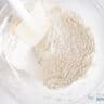 A glass bowl where the dry ingredients for the carrot cake are mixed with a white spatula.