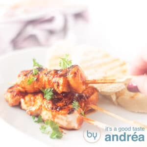 A square photo with a white plate with three chicken skewers, a hand grabbing one and three slices of toast