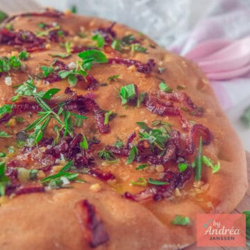 A square photo showing a portion of focaccia topped with fresh herbs, butter, garlic, and fresh herbs.