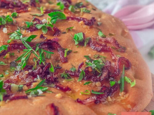 A square photo showing a portion of focaccia topped with fresh herbs, butter, garlic, and fresh herbs.