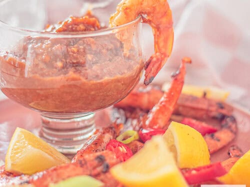 A square photo with a glass filled with cashew satay sauce and grilled shrimps, spring onion, lemon and peppers surrounding it.