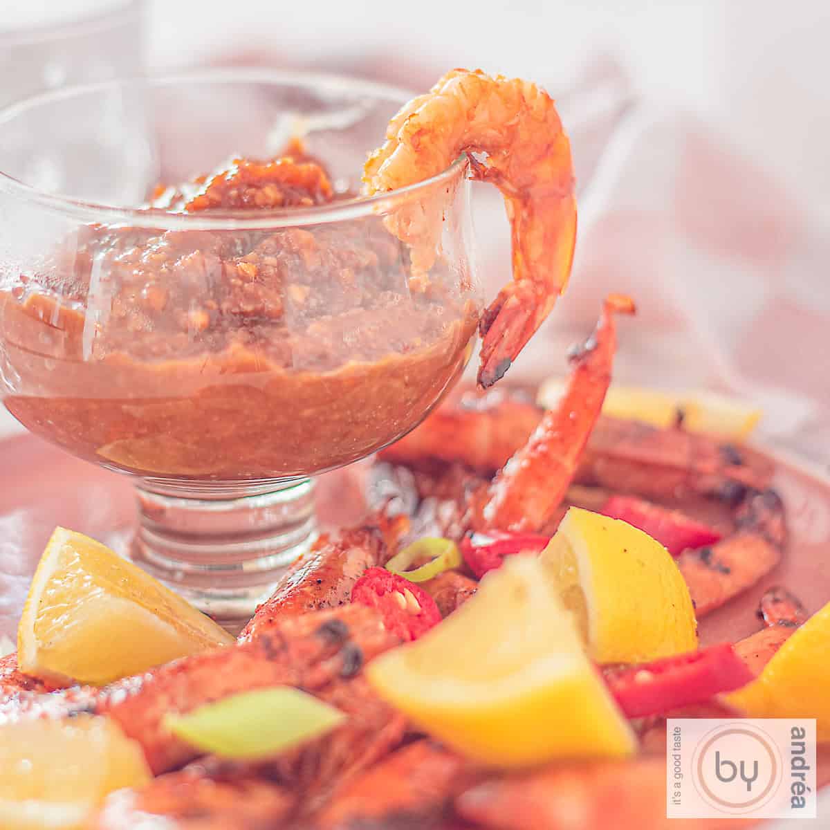 A square photo with a glass filled with cashew satay sauce and grilled shrimps, spring onion, lemon and peppers surrounding it.