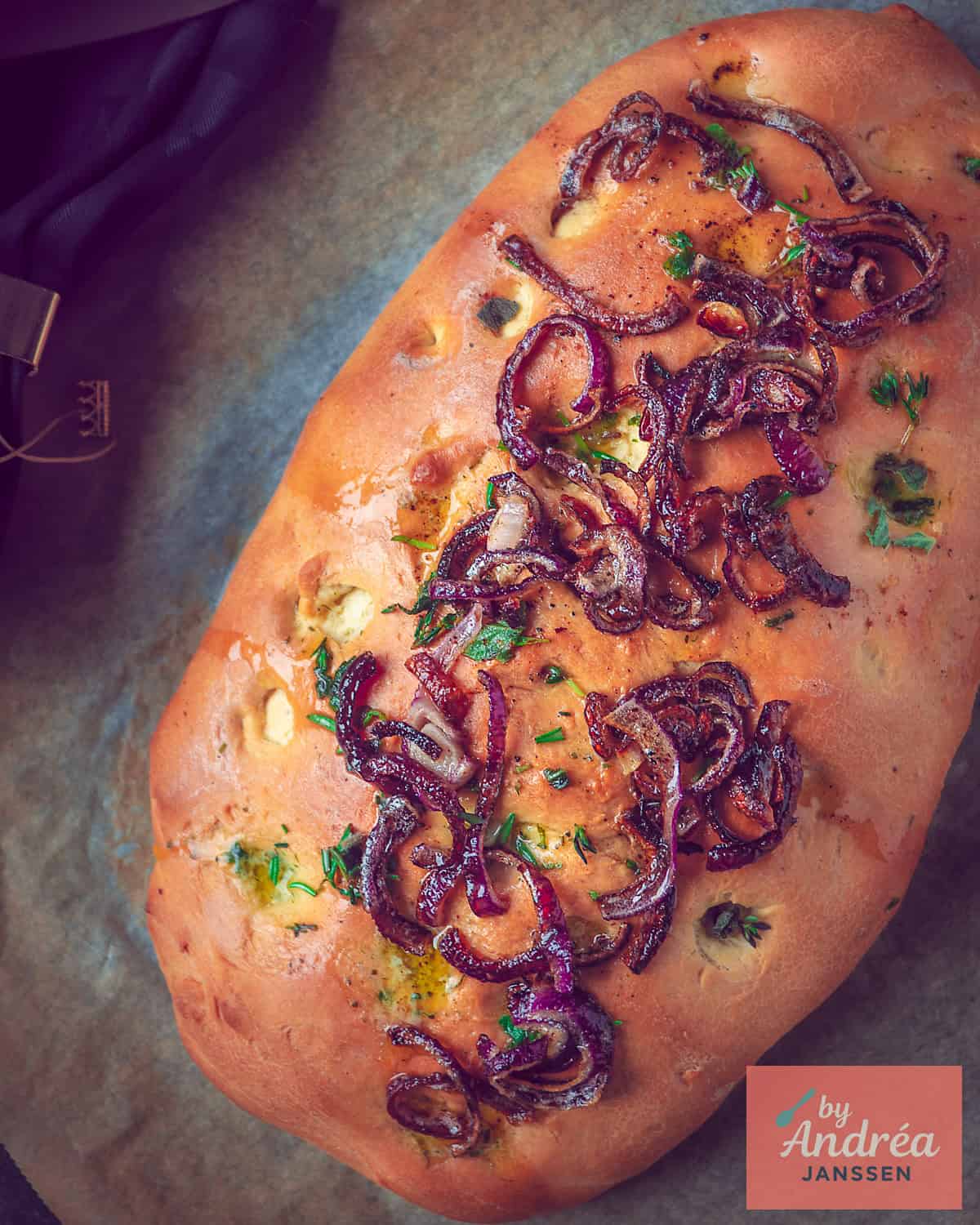 Een foto van bovenaf van een focaccia met rode ui, kruiden en boter op een bakplaat.